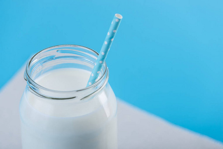 玻璃瓶新鲜的牛奶与稻草在蓝色背景。多彩简约。健康乳制品与钙的概念