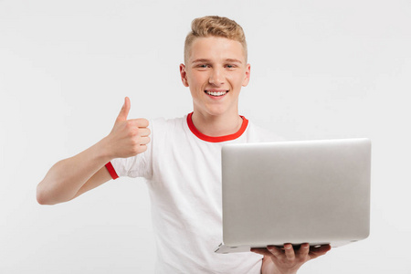 照片的积极年轻男子穿着 t恤衫微笑着, 显示拇指, 而持有银色笔记本电脑隔离在白色背景