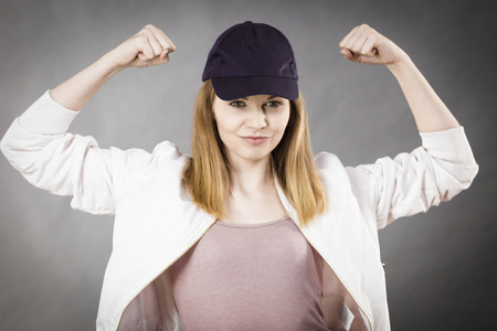 年轻的运动型女人戴帽和运动服显示她手臂的肌肉享受的锻炼效果。工作室拍摄灰色背景