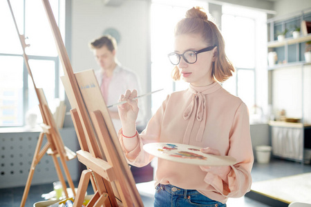漂亮的女孩穿着米色上衣和眼镜站在画架与调色板和绘画的东西与画笔和油颜色