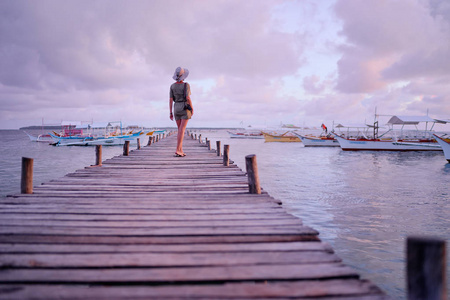 戴着帽子的年轻女子手持相机站在木制的钓鱼码头上, 拥有美丽的热带海景。