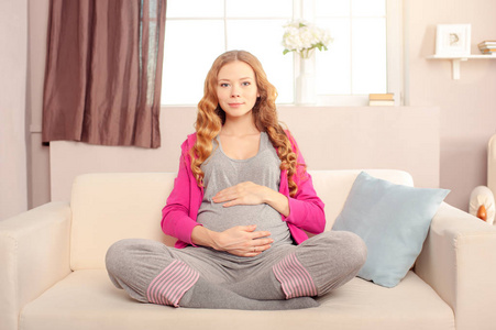 美丽的孕妇坐在莲花位置在沙发上和微笑