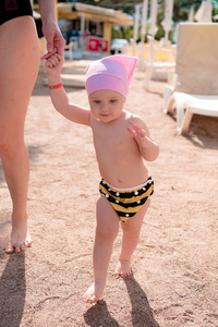女孩与她的母亲手拉手在一个公共海滩上的大沙滩上, 在沙滩躺椅之间, 她穿着粉红色的帽子和条纹游泳内裤