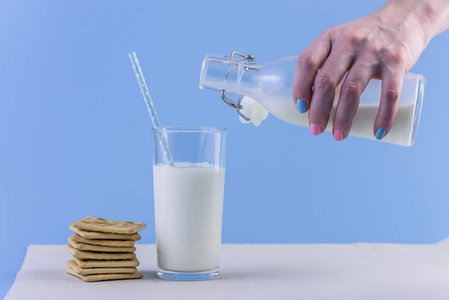 一个女人的手把新鲜的牛奶从玻璃瓶倒入玻璃和蓝色背景的饼干。多彩简约。健康乳制品与钙的概念