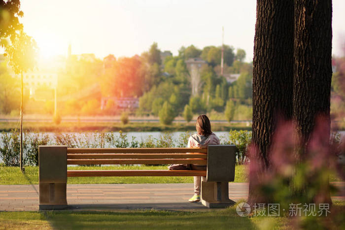 孤独的年轻女子在湖附近的公园里的长椅上坐在日落