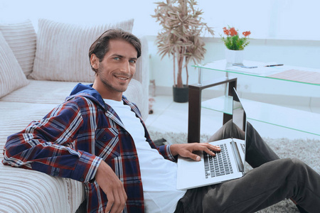 年轻的男人微笑着坐在沙发边的地板上的笔记本电脑