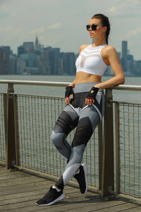 美丽的年轻健身模型穿着时髦的运动服摆在码头与城市背景