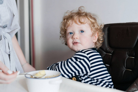 可爱的卷发蓝眼睛的男孩坐在厨房里, 妈妈用健康的食物喂养他。