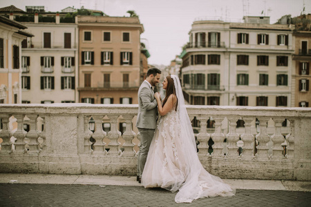 年轻迷人新婚夫妇步行和摆在罗马与美丽的古建筑在他们的婚礼当天的背景
