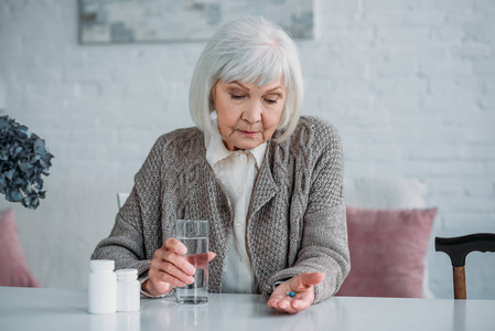 灰色头发妇女的画像用药丸和杯子水在手单独坐在桌在家