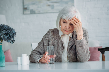 灰色头发妇女的画像坐在桌用药片和玻璃水在家