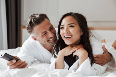 照片的多民族夫妇白人男子和亚洲妇女穿着白色浴衣躺在家里的卧室或酒店公寓与遥控器在手看电视