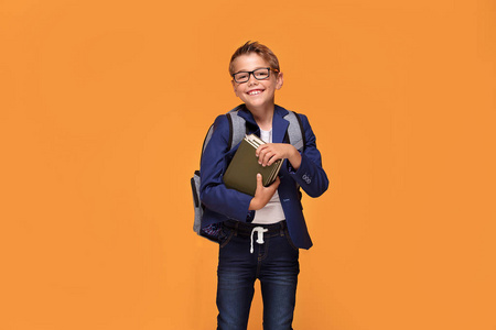 小男孩在眼镜与背包站立在橙色背景, 拿着书, 微笑