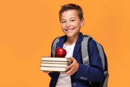 小学校男孩与背包站在橙色背景, 拿着书和苹果, 微笑着