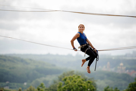 侧面视图微笑金发女郎坐在 slackline 绳子上的房子在树和晴朗的天空的背景下