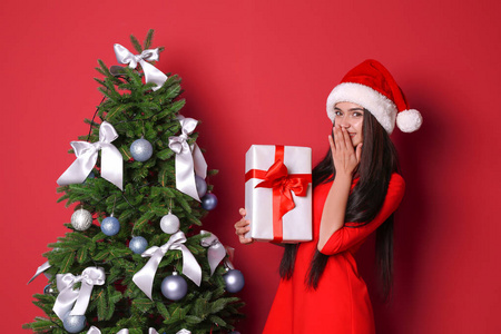 美丽的年轻女子在圣诞老人的帽子与礼品盒附近的圣诞树上的颜色背景