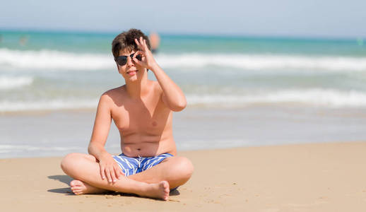 小孩子在假日在海滩与愉快的面孔微笑做 ok 标志用手在眼睛看通过手指