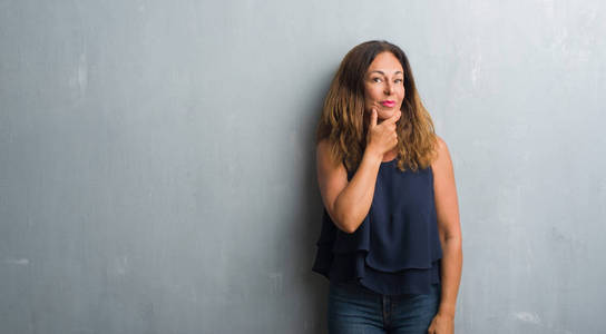 中年拉美裔妇女站在灰色的垃圾墙上, 看着自信的相机与交叉的手臂和手举起下巴。积极思考