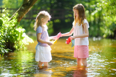 两个小妹妹在温暖晴朗的夏日里在河边玩纸船。孩子们在水里玩得很开心。幼儿暑期活动