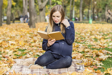 年轻的怀孕女孩读了一本书, 坐在一个秋季公园的格子。秋季和户外休闲的概念