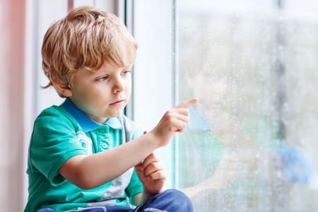 金发碧眼的小孩男孩坐在窗户旁边，看雨滴