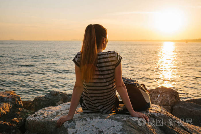 年轻游客的女孩背着日落时分坐在海边岩石上,眺望远方