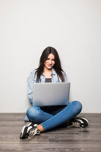 快乐的年轻女子坐在地板上与交叉的双腿和在灰色的背景上使用笔记本电脑