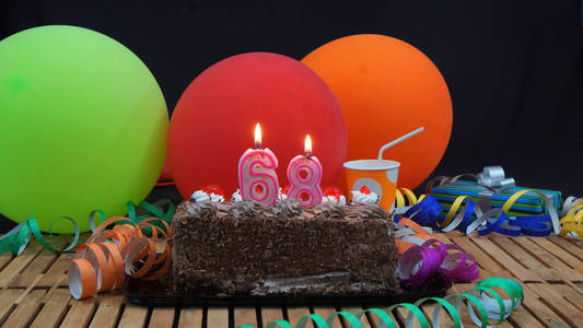 用蜡烛燃烧与五颜六色的气球 礼品 塑料杯和飘带与黑色背景背景仿古木制的桌子上的巧克力生日蛋糕