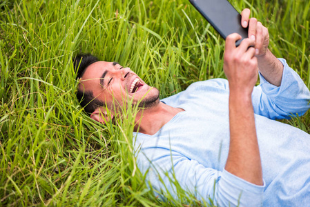 年轻的快乐商人微笑和躺在绿色的草地，在设备上看