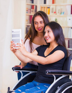 年轻的女人坐在轮椅使用手机与助理站在她旁边，两个微笑愉快地表现出积极的态度，体能恢复概念