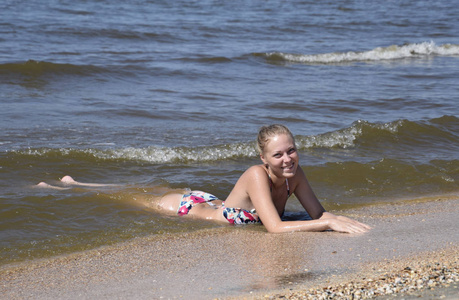 金发女孩穿着比基尼躺在沙滩和海浪溅上它。美丽的年轻女子在海面背景的彩色比基尼