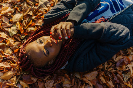 美丽的非洲裔美国女孩躺在黄色的叶子在秋天的公园。女性制作光谱分析中的应用和微笑