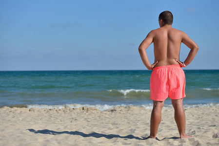 健壮的年轻男人在粉红色短裤坐在沙滩上，看着