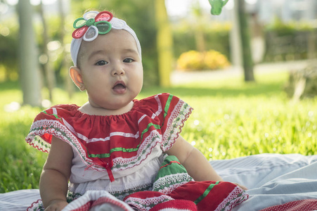 宝贝很漂亮白色，坐在草地上，墨西哥的彩色的连衣裙，戴着白丝带和鞠躬领带的头上。打着手势，表达不同的情绪，原来和非常友好而风趣，暗