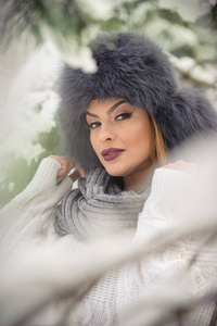 白色套衫与尺寸过大的毛皮帽享受森林的冬季景色美丽的女人。白雪覆盖的树木树枝下构成的金发女孩。有吸引力的年轻女性，在明亮的冷的天，