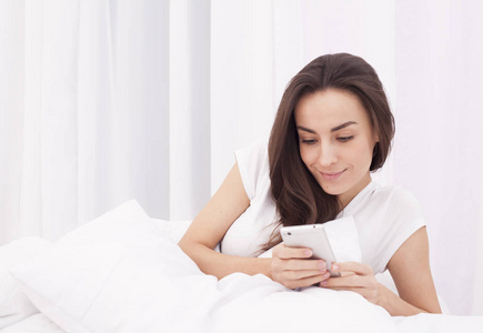 很漂亮年轻的黑发女人将消息写入手机在白色的床上