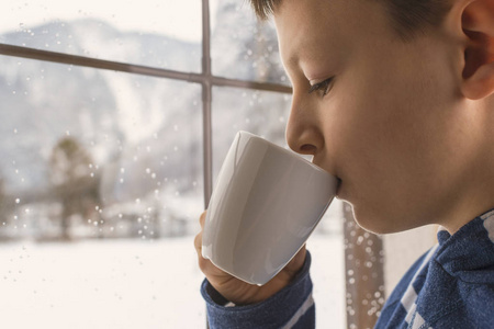茶杯在手望着窗外没有在冬天雪山地森林景观的忧郁男孩