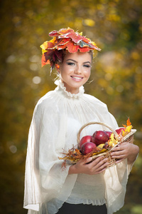 秋天的女人。 户外拍摄中美丽的创意妆容和发型。 头发上有叶子的女孩拿着一个带苹果的篮子