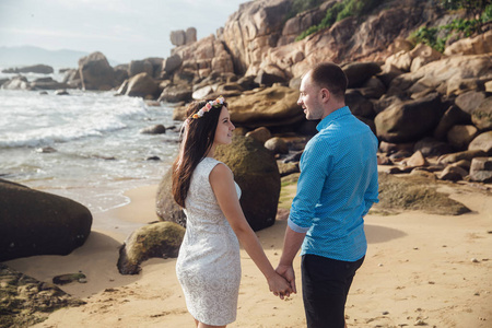 在爱拥抱 牵手海的海滩上夏天的年轻夫妇。那个穿着蓝色的衬衫和花圈在她的头发穿白裙子的女孩