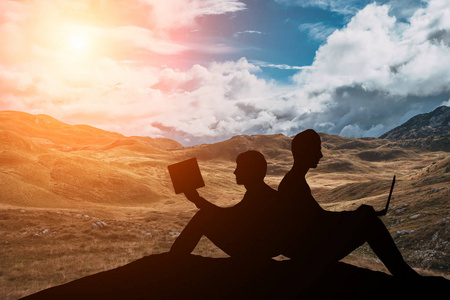 两个朋友坐在读书的山上, 坐在美丽日落的背景下的笔记本电脑上