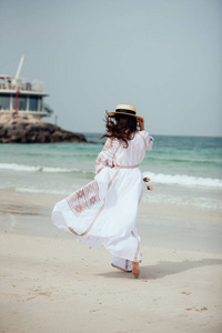 美丽的年轻女子穿着白色的长裙，长发和帽子沿着海滩走
