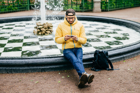 黑帽子 眼镜 牛仔裤和黄色夹克坐在公园玩游戏在休息后长漂移通过市欣赏新鲜空气他手机上的喷泉附近的时髦旅行家