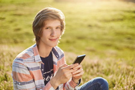 高兴的小男孩，与狭窄的黑眼睛和时尚的头发坐在草地上拿手机检查他的电子邮件或使用免费的互联网连接，而停留在美好的绿色领域的新闻阅读