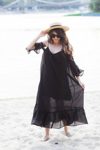 美丽年轻的黑发女人，在黑色的连衣裙，黑色的太阳镜和草帽走在海滩上的沙，并构成在夏季日出