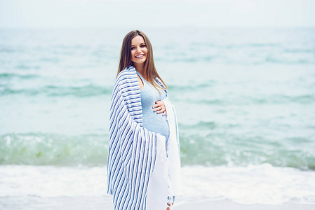 美丽孕妇穿着蓝色紧身连衣裙布满白色条纹格子上海滩摸她的肚子爱与关怀。走在海边，在阳光灿烂的日子