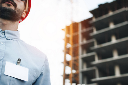 空白的白色徽章或胸部的一件衬衫的现代工程师或建筑师在建筑工地为背景的红色安全帽的商业名片。年轻商人与空白白色徽章佩在胸前的衬衣