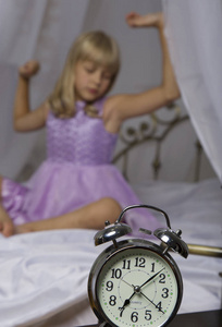 闹钟站在床边的桌子上。醒来的一个熟睡的年轻女孩是伸展在床上的背景