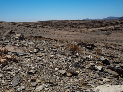 艰苦生活的复制空间显示了纳米布沙漠的岩石山干燥尘土飞扬的景观地面与分裂页岩块, 其他石头, 沙漠植物和蓝天