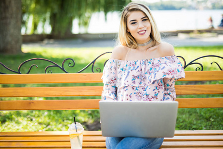 美丽的年轻金发碧眼女学生坐在板凳上使用便携式笔记本电脑。年轻漂亮的女孩使用个人便携式计算机在因特网上查找信息