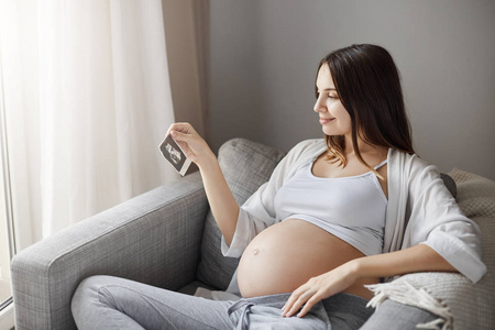 怀孕女士看着婴儿超声图片试图猜测是男孩还是女孩。在她舒适的沙发里呆在家里